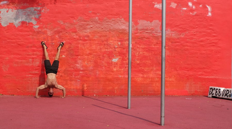 Full length of shirtless man doing handstand against red wall