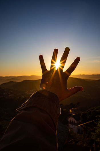 Cropped hand of person against mountains and sky during sunset