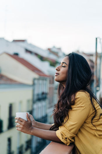 Side view of young hispanic woman with closed eyes enjoying hot beverage while resting on balcony in evening