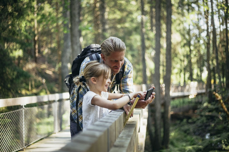 Smiling father with backpack and daughter looking at smart phone in forest