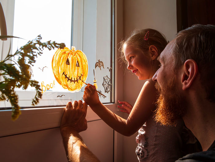 Child father painting pumpkin on window preparing halloween little girl dad drawing decorating room