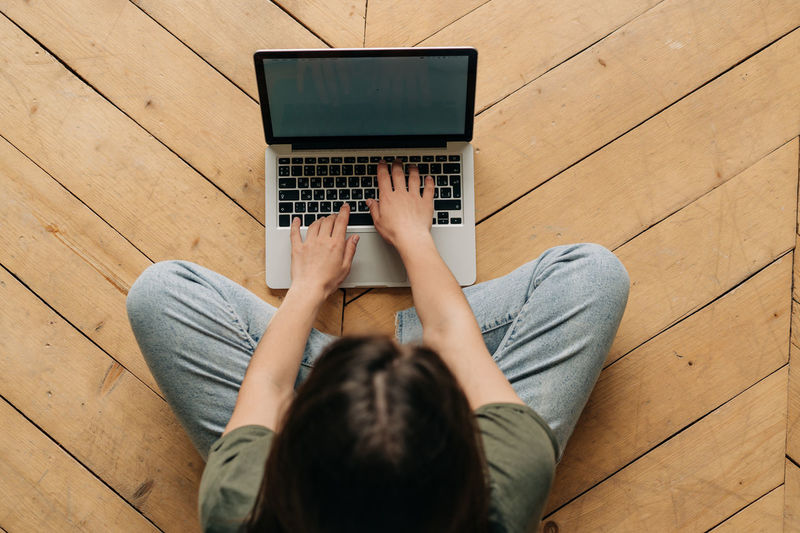 Top view of an unrecognizable woman working on a laptop while sitting on the floor.