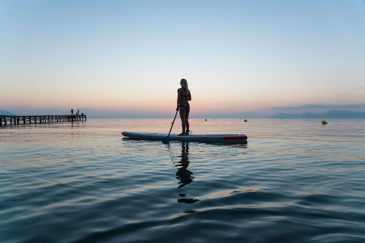 Back view of silhouette of unrecognizable female surfer standing on paddleboard and rowing against spectacular sun in sunset sky