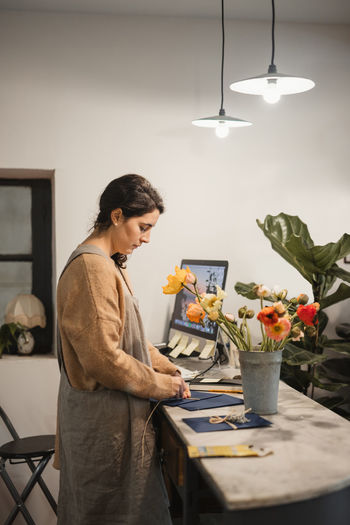 Diligent worker in brown sweater and gray apron concentrating and making decor elements for designing modern apartments while standing at table with computer and fresh peach color flowers in vase in contemporary flower shop