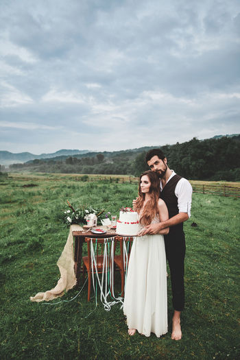 Full length of young couple holding wedding cake while standing on field against cloudy sky