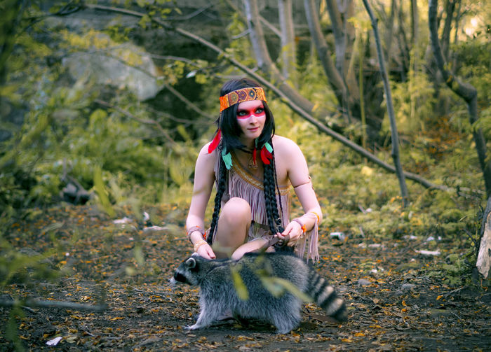 Young woman in traditional clothing looking away while crouching by raccoon in forest