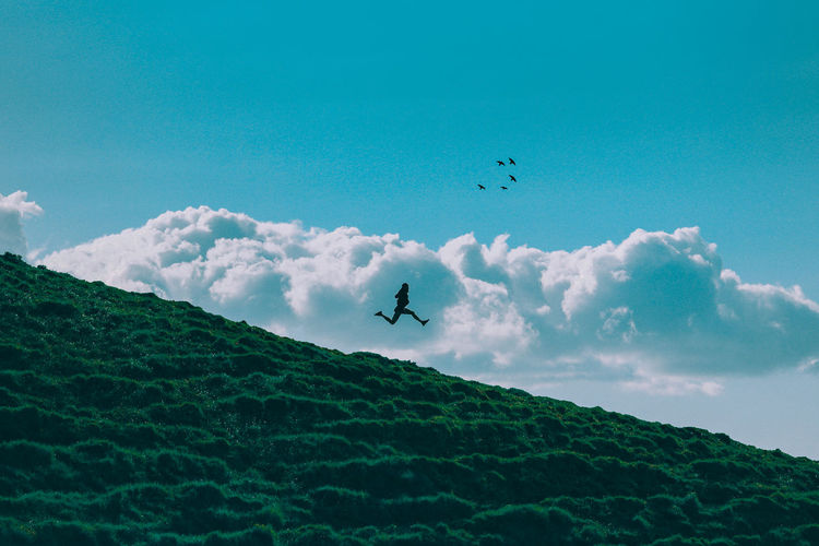 Silhouette hiker jumping over hill against blue sky