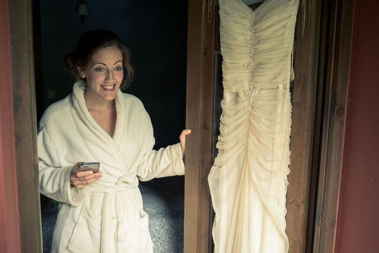 Smiling bride in bathrobe standing wedding dress on door in bedroom