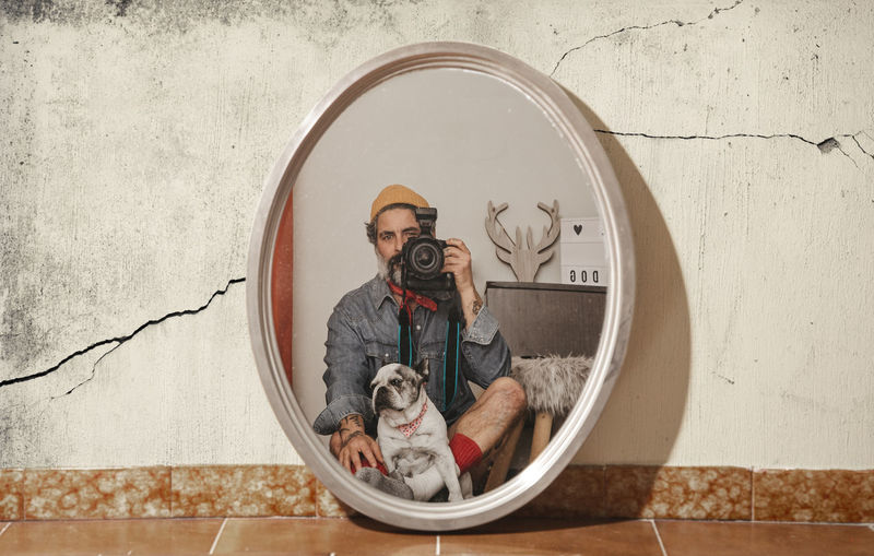 Portrait of man clicking photograph with dog while looking at mirror
