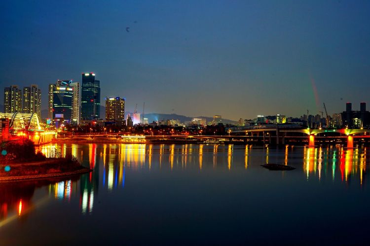 Illuminated buildings by river against sky at night