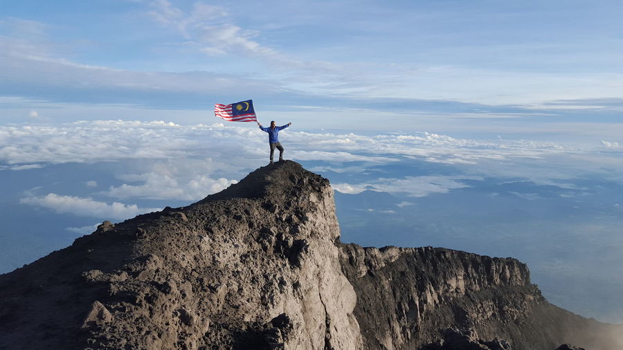Man holding malaysian flag while standing on mountain against cloudy sky