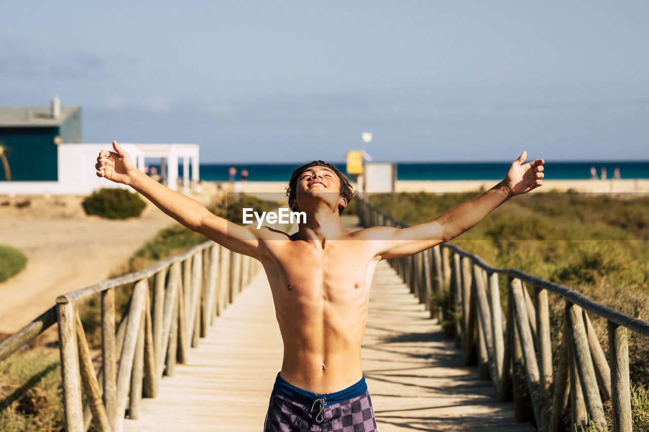 MIDSECTION OF SHIRTLESS MAN STANDING ON RAILING AGAINST SKY