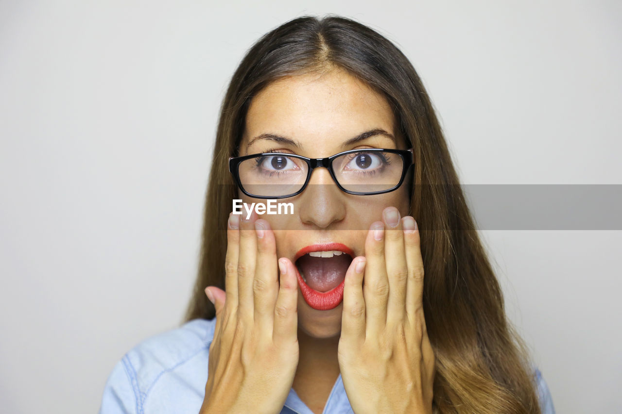 Close-up portrait of shocked young woman wearing eyeglasses against gray background