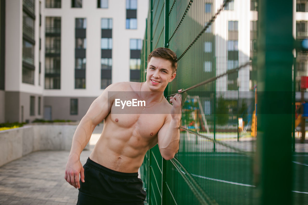 Portrait of muscular man standing by fence at park