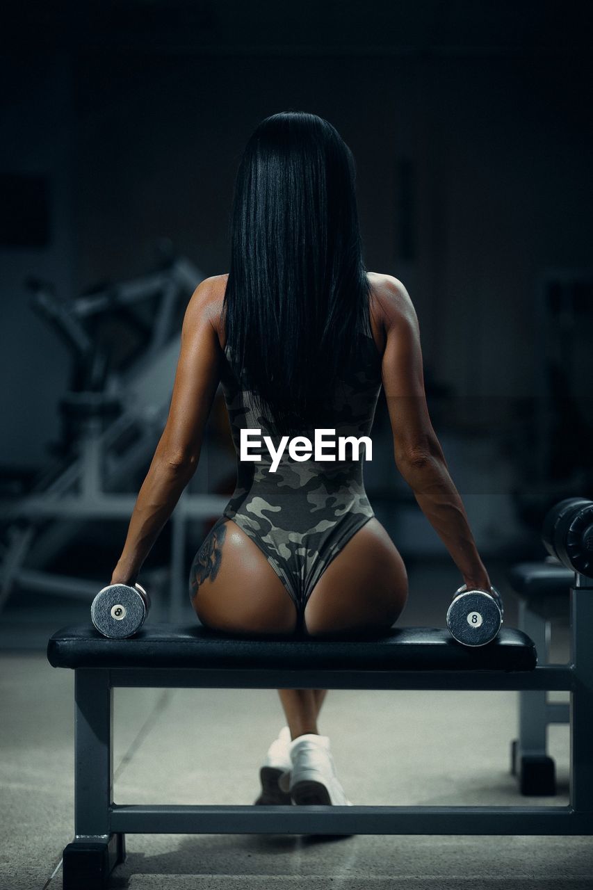 Rear view of woman sitting with dumbbells on bench in gym