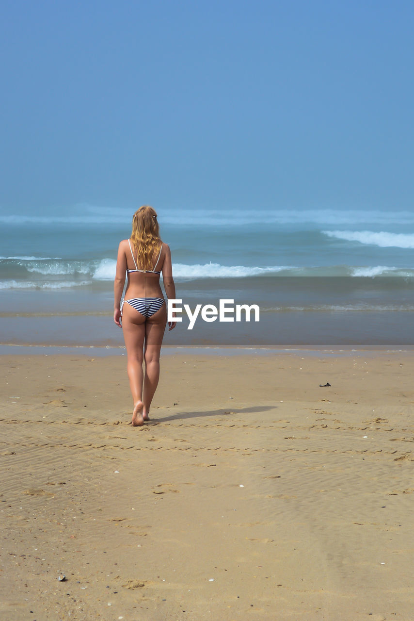 Rear view of woman wearing bikini while walking at beach against clear sky