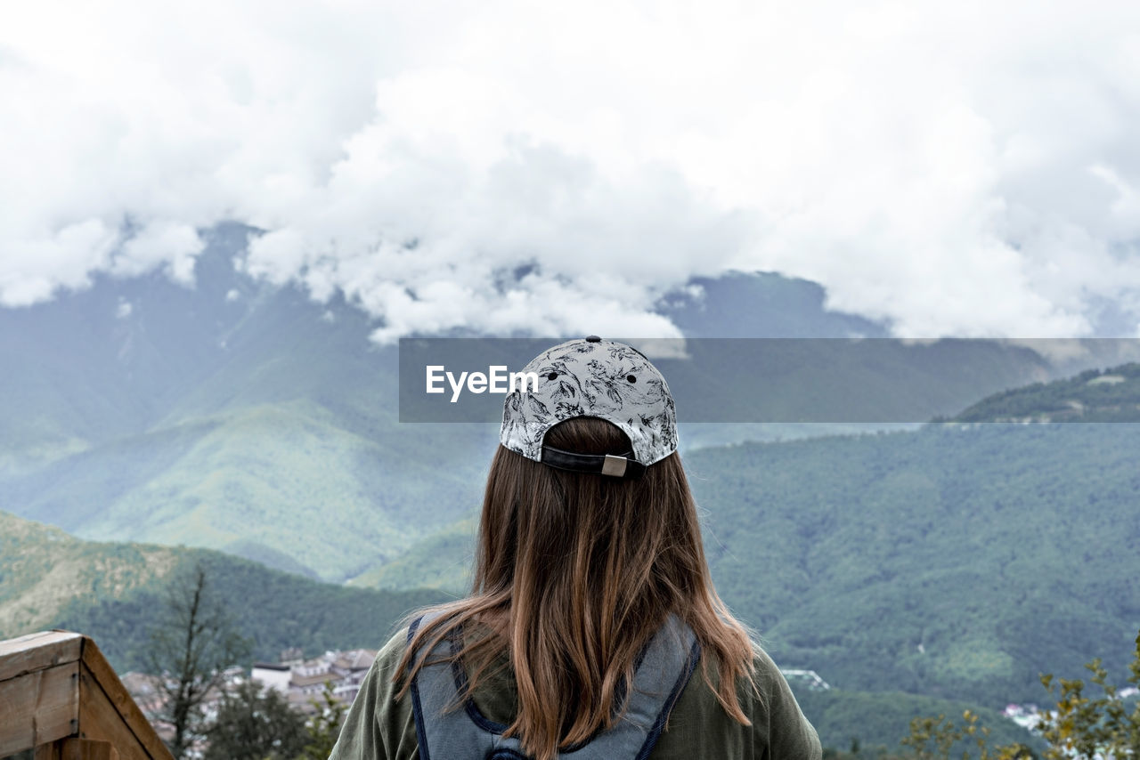 Rear view of woman in cap with backpack looking at view of mountains and cloudy sky on  viewpoint