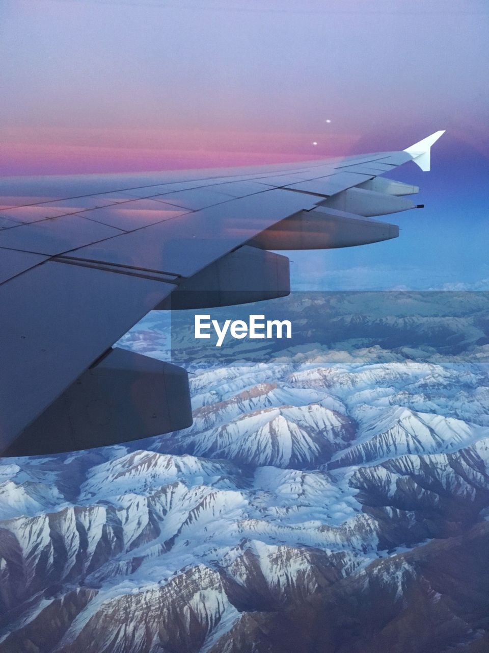 Cropped image of airplane flying over snow covered mountains against sky during sunset
