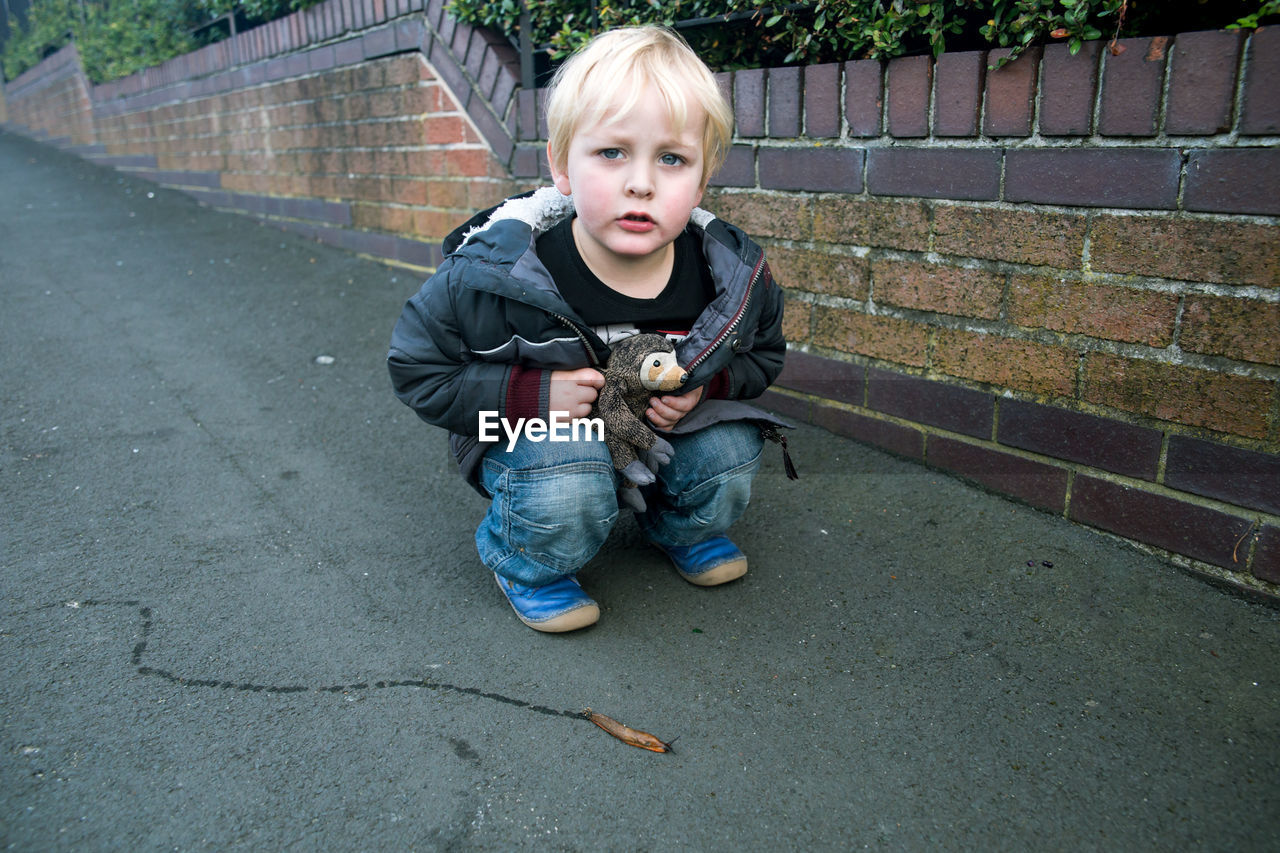 Portrait of boy with stuffed toy crouching by slug on street against surrounding wall