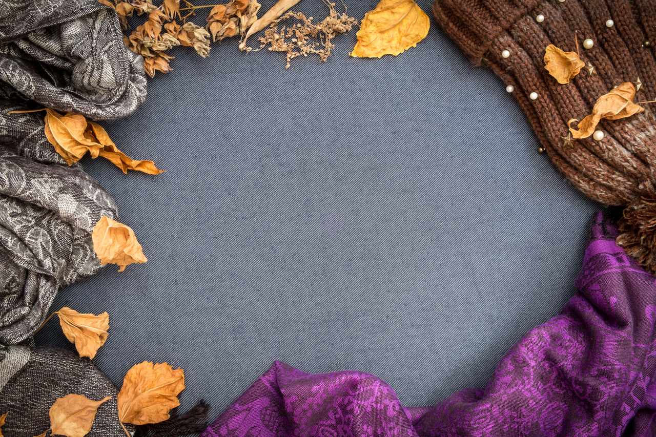 Autumn winter flat lay. brown knitted beanie, purple scarf and dry leaves on blue blank background.