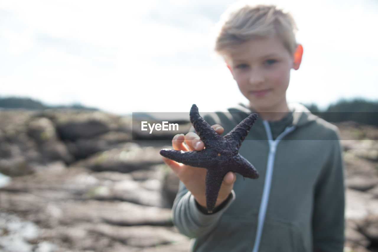 Portrait of boy holding starfish at beach against sky