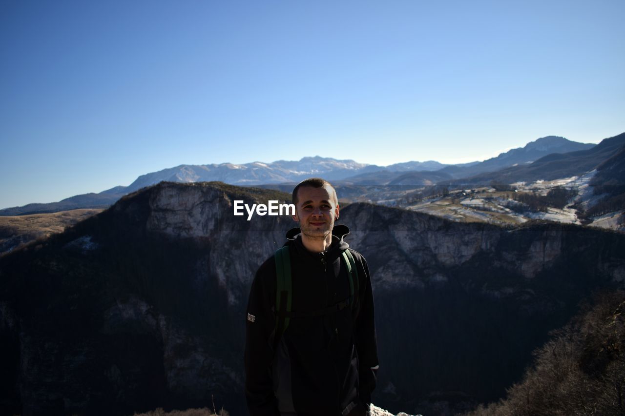 PORTRAIT OF SMILING YOUNG WOMAN STANDING AGAINST MOUNTAINS