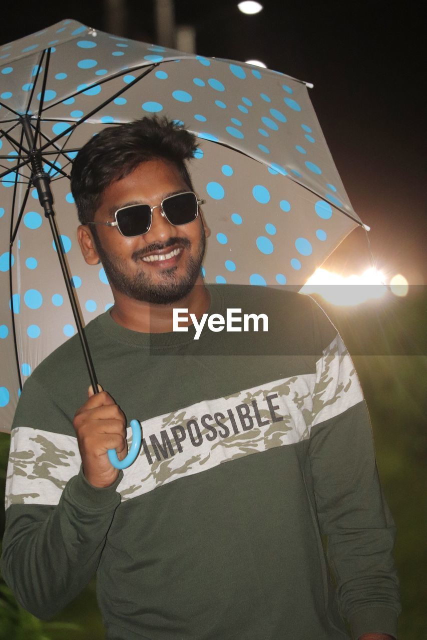 umbrella, fashion, one person, sunglasses, glasses, protection, adult, men, holding, portrait, person, security, young adult, waist up, front view, clothing, human face, smiling, happiness, night, looking at camera, fashion accessory, emotion, casual clothing, beard, cool attitude, standing, facial hair, arts culture and entertainment, leisure activity, nature