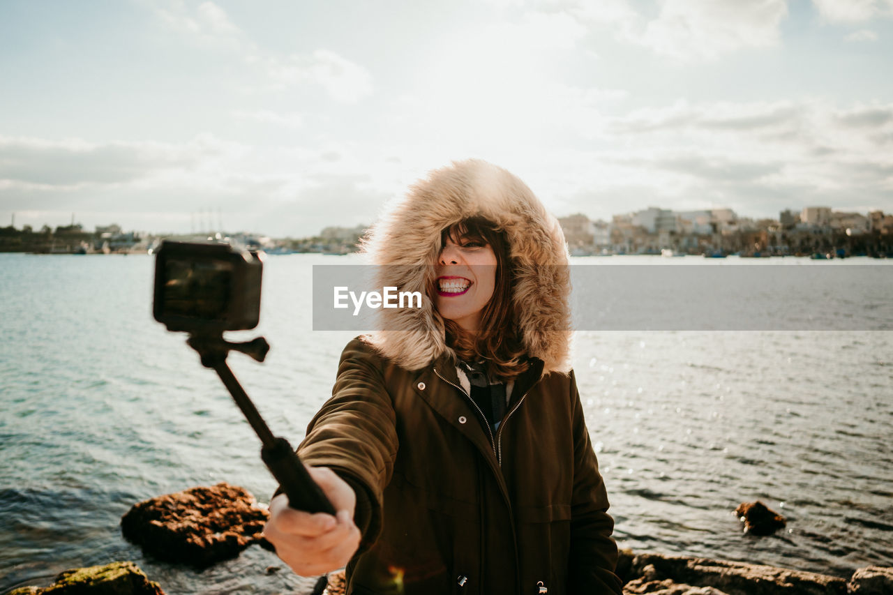 Smiling woman taking selfie with camera while standing against lake