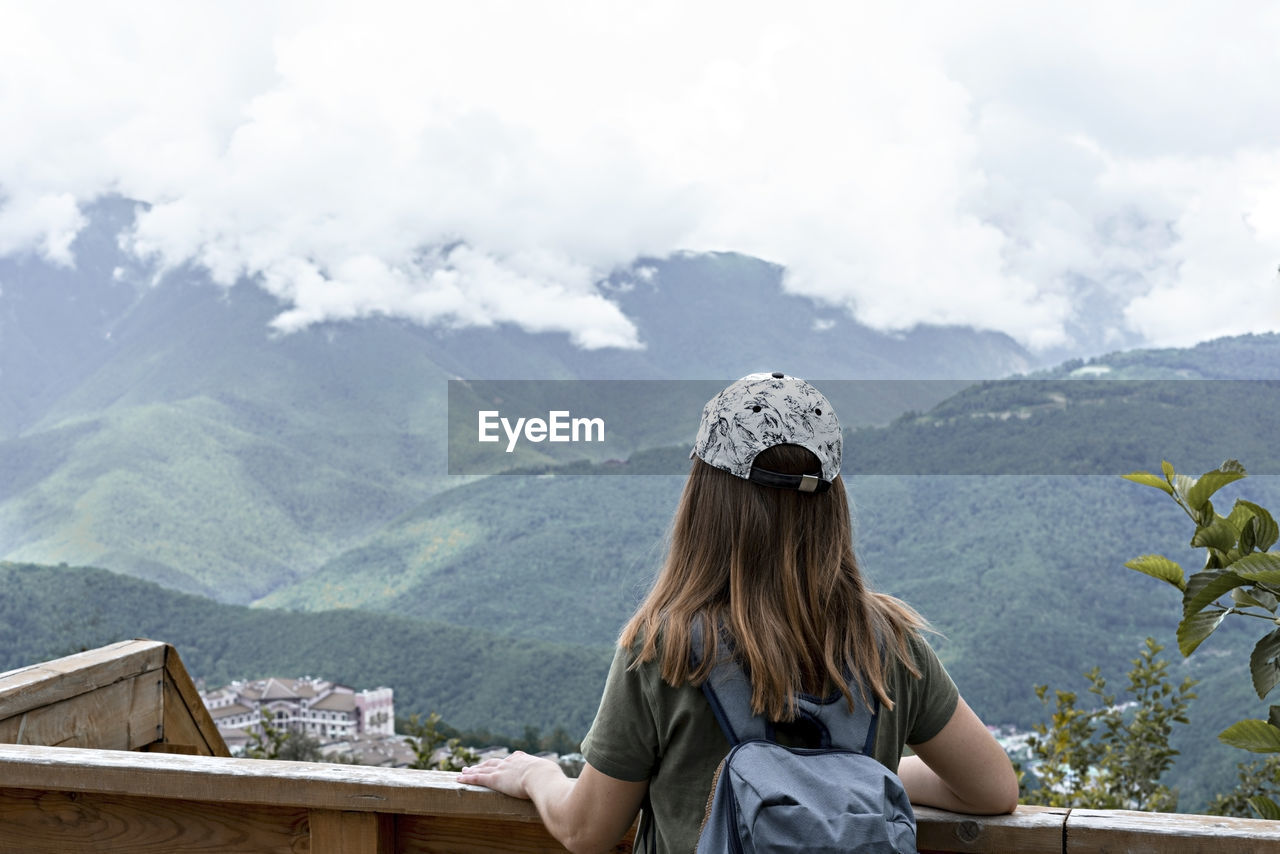Rear view of young woman in cap with backpack looking at view of caucasian mountains and cloudy sky 