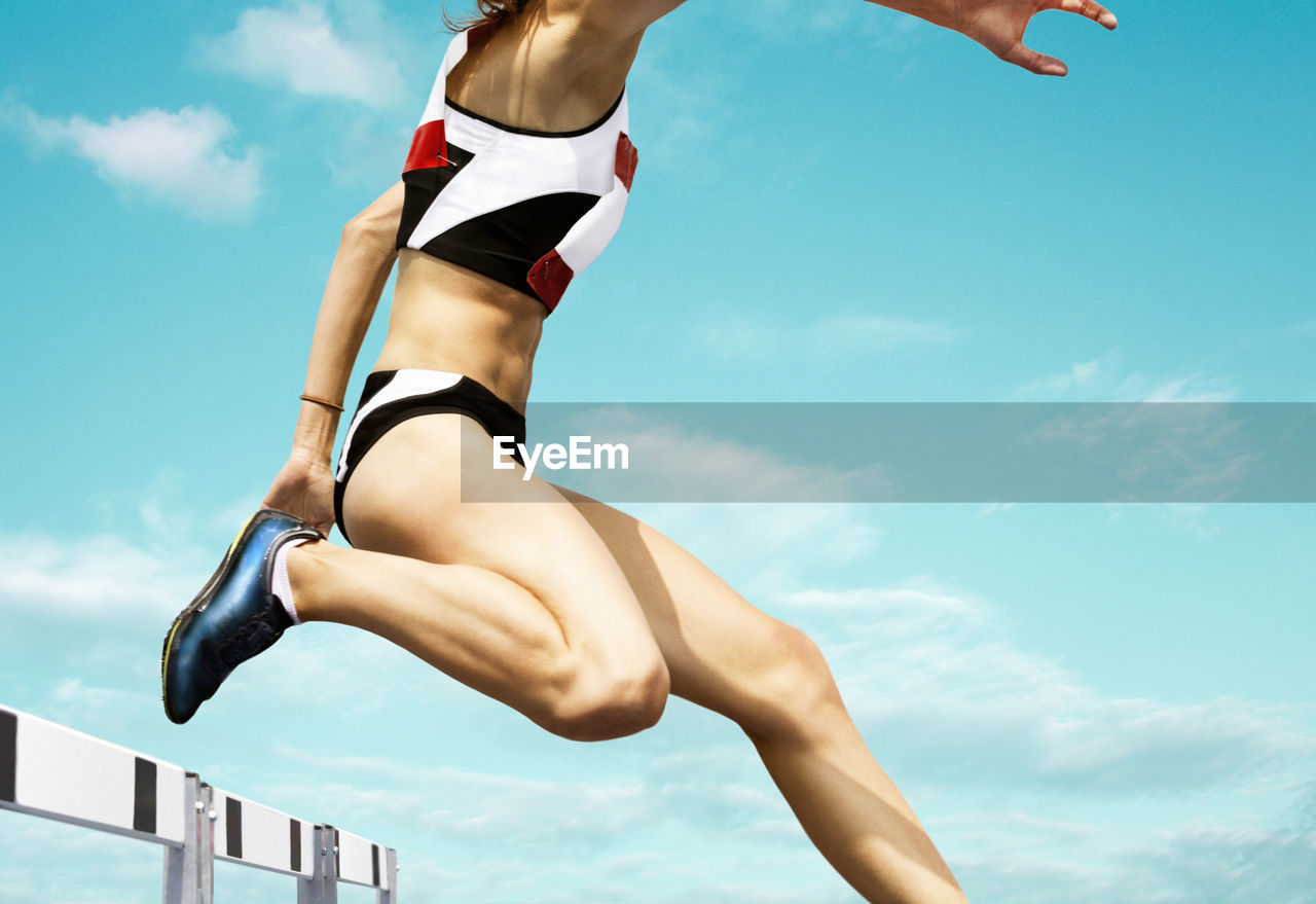 Low section of woman jumping over hurdle against blue sky
