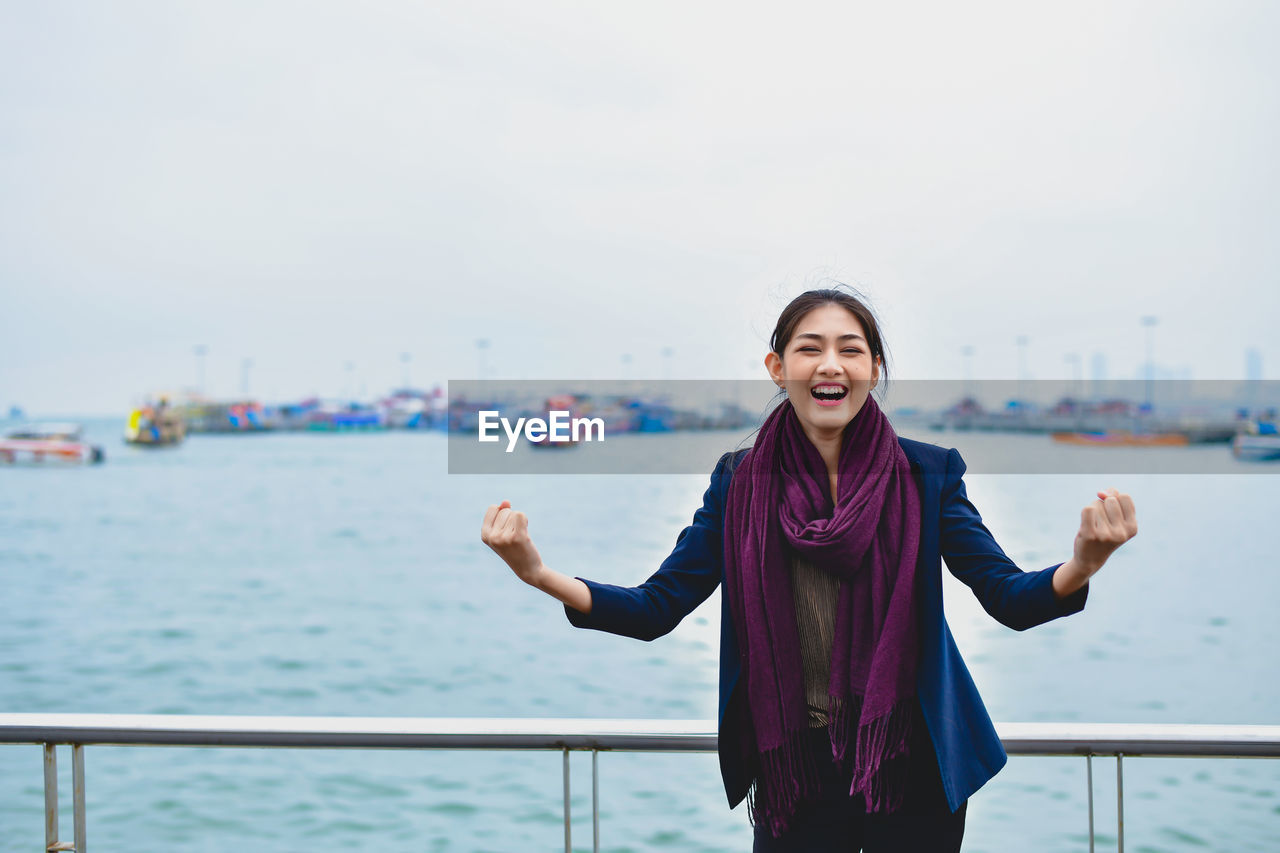 Cheerful young woman clenching fists while standing against sea