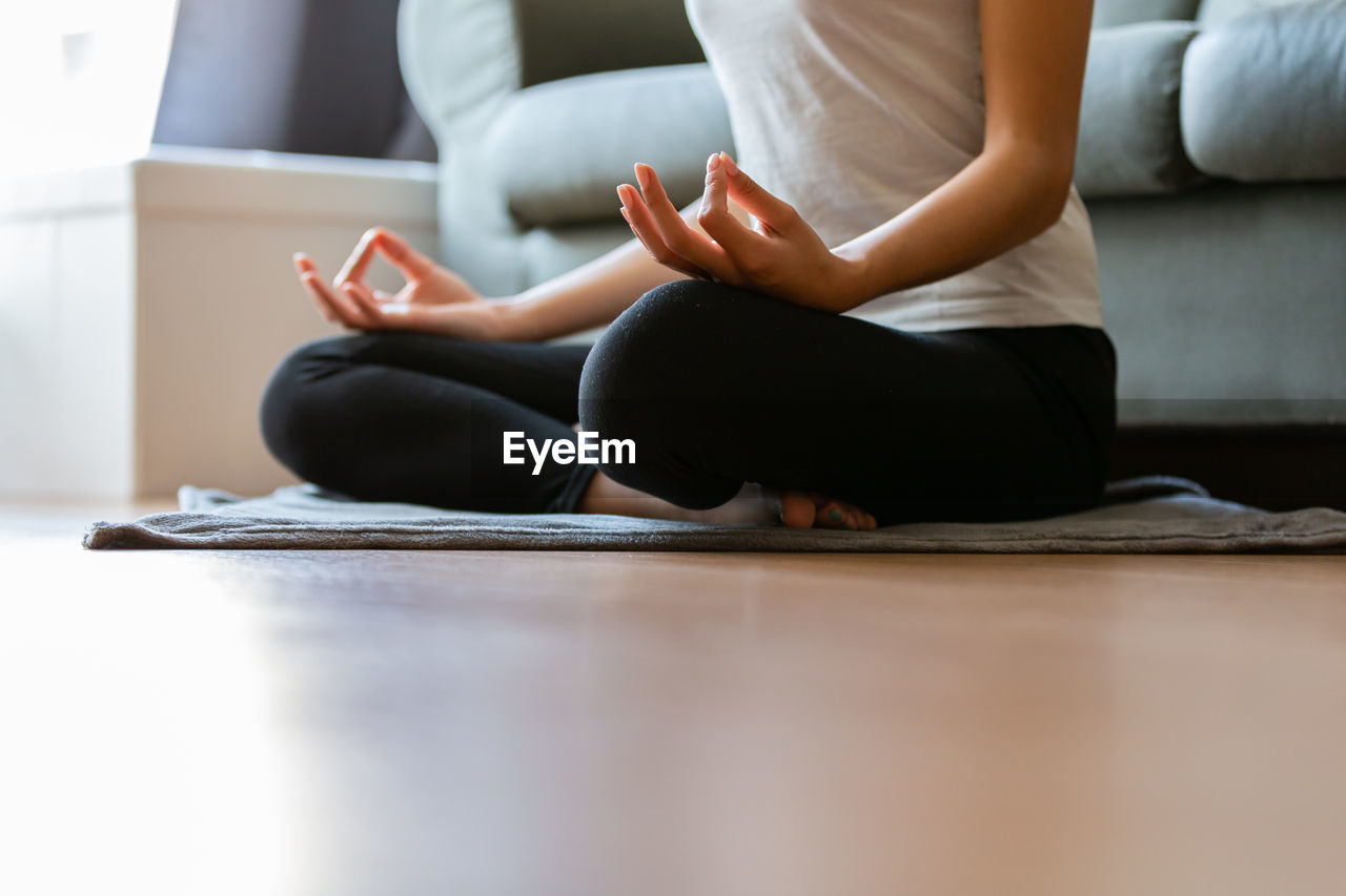 sitting, adult, yoga, lifestyles, indoors, physical fitness, women, exercising, one person, relaxation, wellbeing, cross-legged, sports, zen-like, meditating, female, sports clothing, barefoot, limb, posture, domestic room, spirituality, stretching, full length, flooring, flexibility, young adult, arm, home interior, exercise mat, tranquility, living room, clothing, human leg, leisure activity, sitting on floor, lotus position, domestic life, practicing, copy space, contact sport, day, pants, martial arts, furniture, concentration, gym, balance, religion, side view