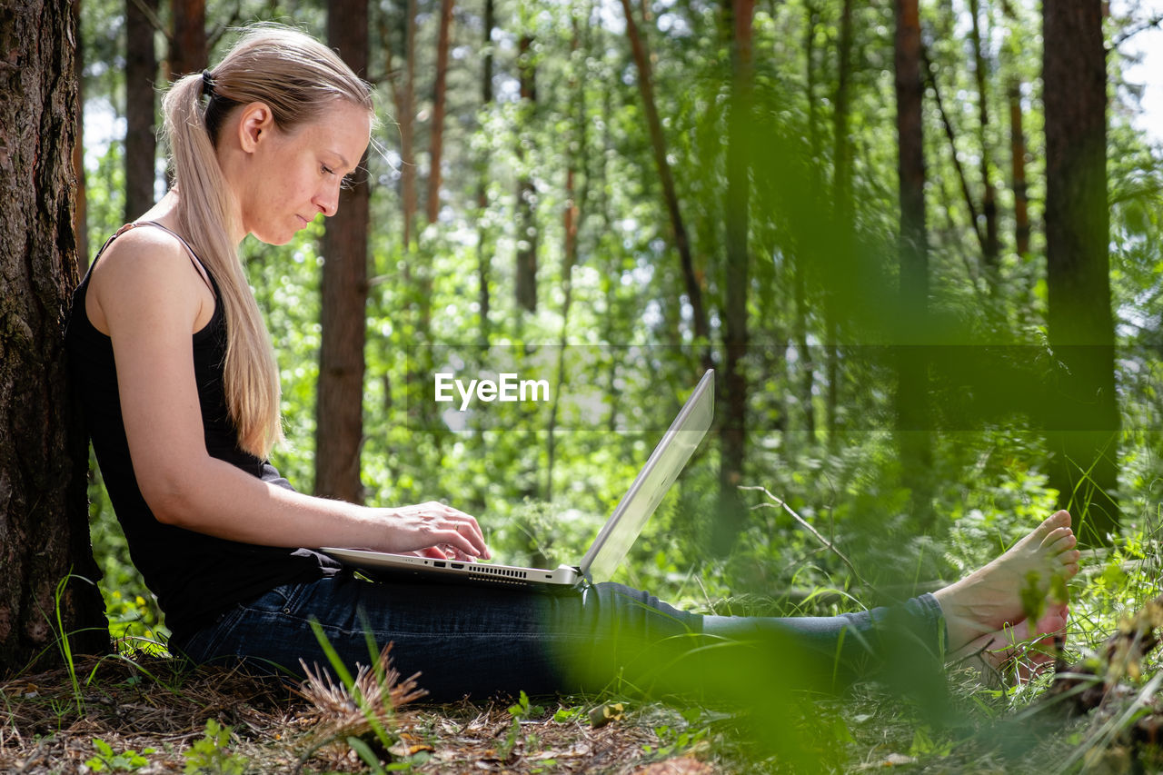 Girl in the forest sits leaning against a tree, working on a laptop. rest at nature. mental health