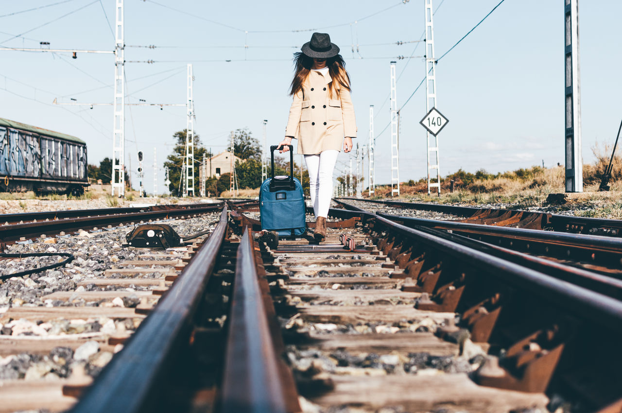 Woman dressed with a grey hat and beige jacket walking with her suitcase along a railway track.