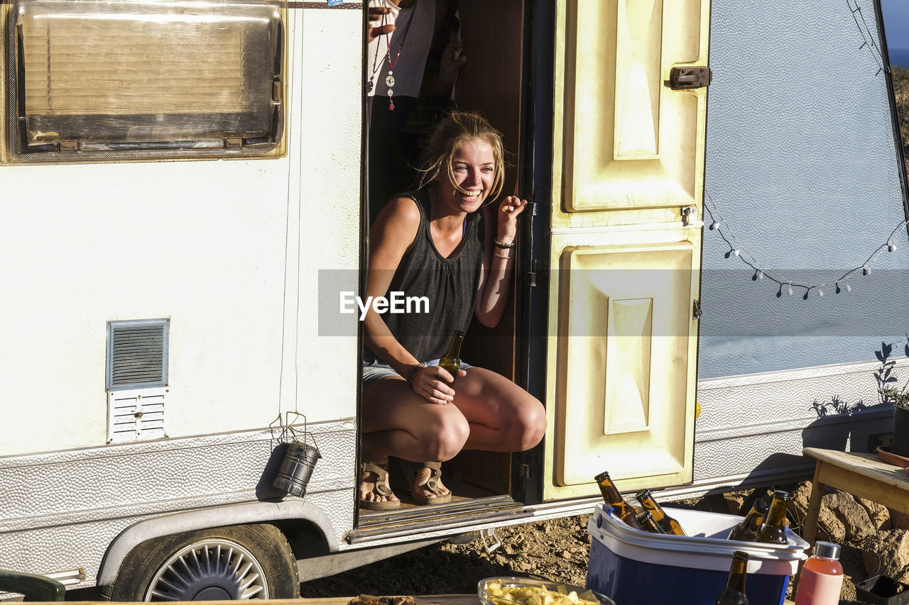 Smiling young woman holding beer bottle while crouching in van