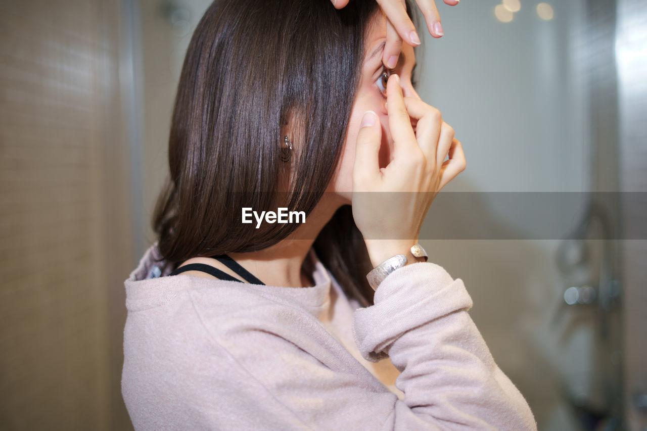 Side view of female putting on contact lens on eye while standing in bathroom and looking in mirror