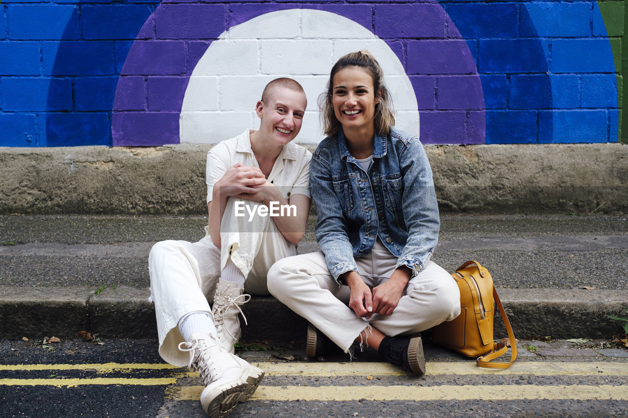 Happy young woman and non-binary person sitting on street