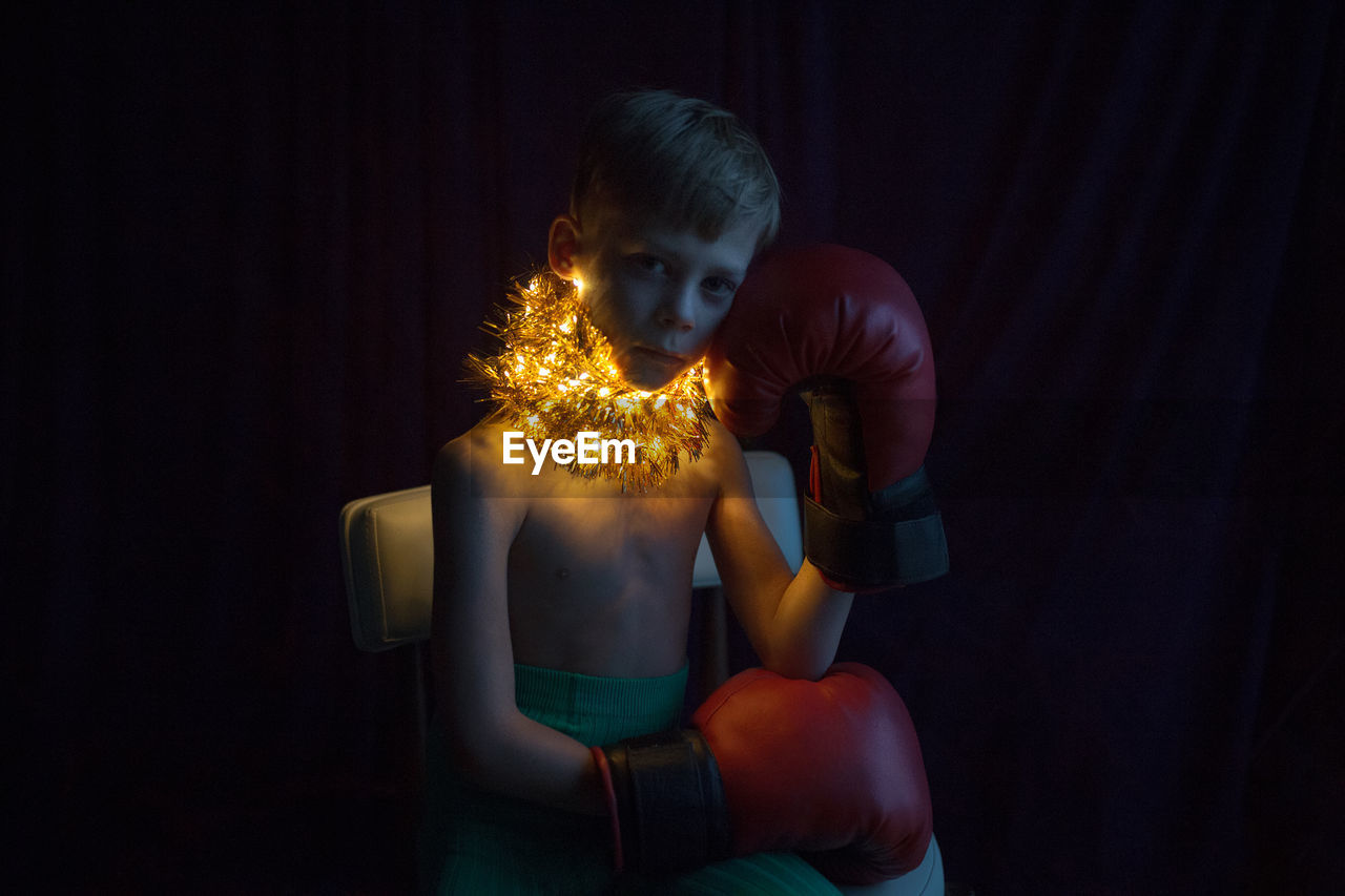 Portrait of shirtless boy with boxing gloves and illuminated lights around neck at home