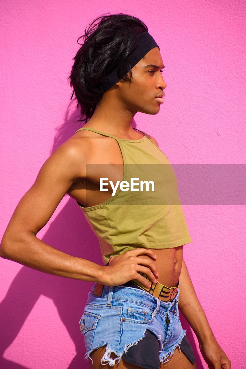 Side view of young model with hand on hip standing against pink wall