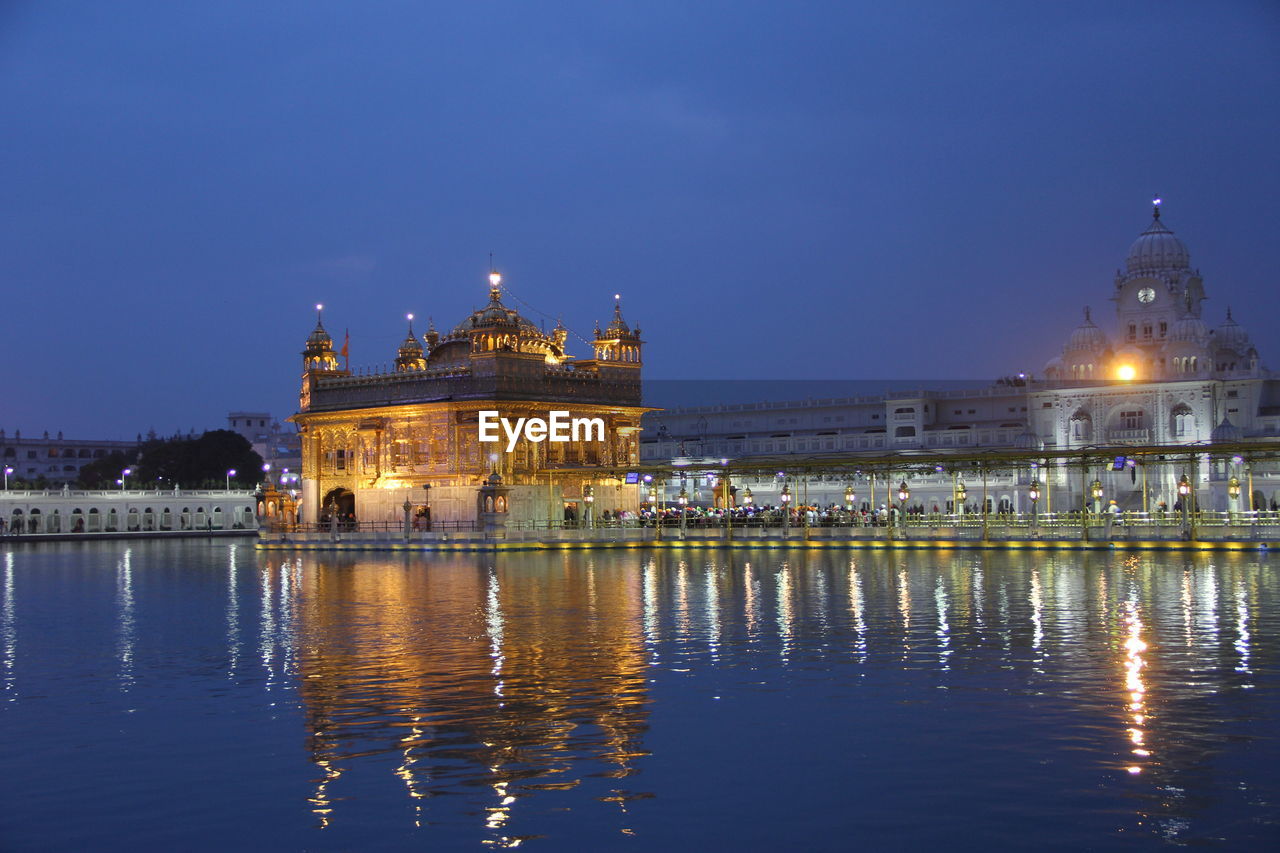 Illuminated golden temple by lake against sky at dusk