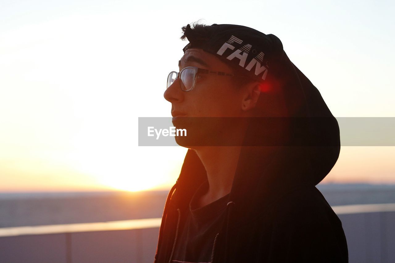 Thoughtful young man in hooded shirt standing against sky during sunset