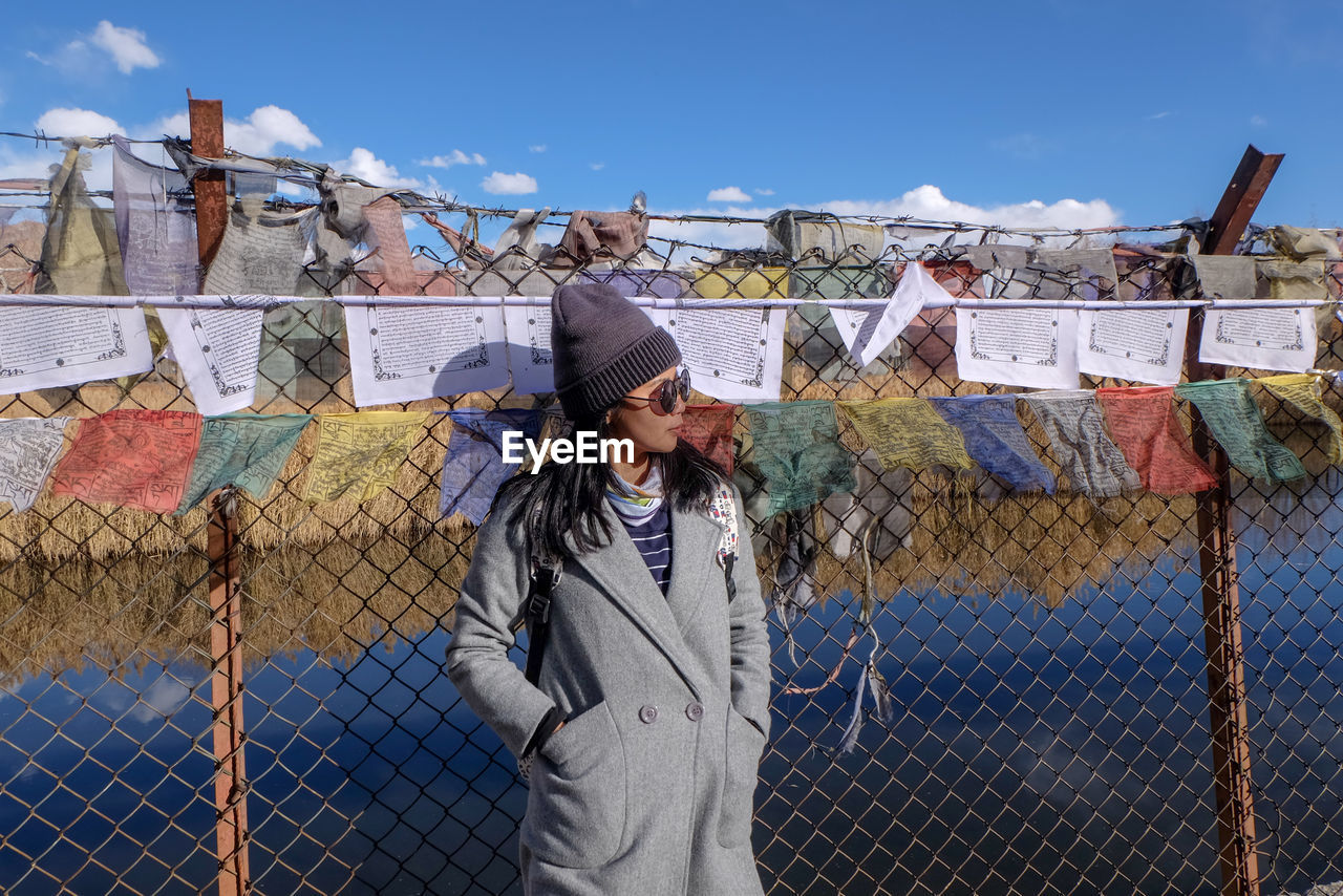 Woman wearing warm clothing while standing against fence and prayer flags