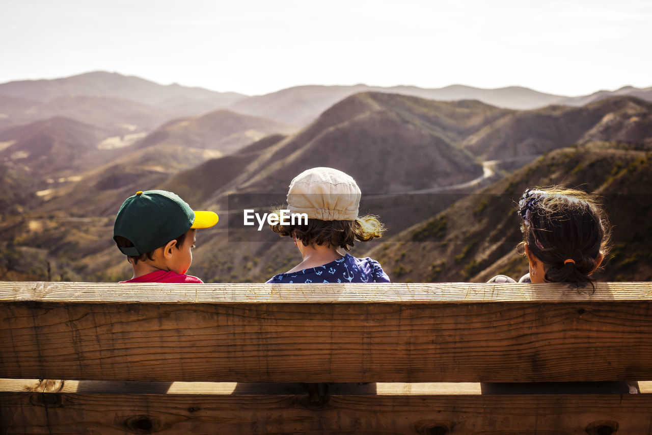 Rear view of siblings sitting on bench against mountains