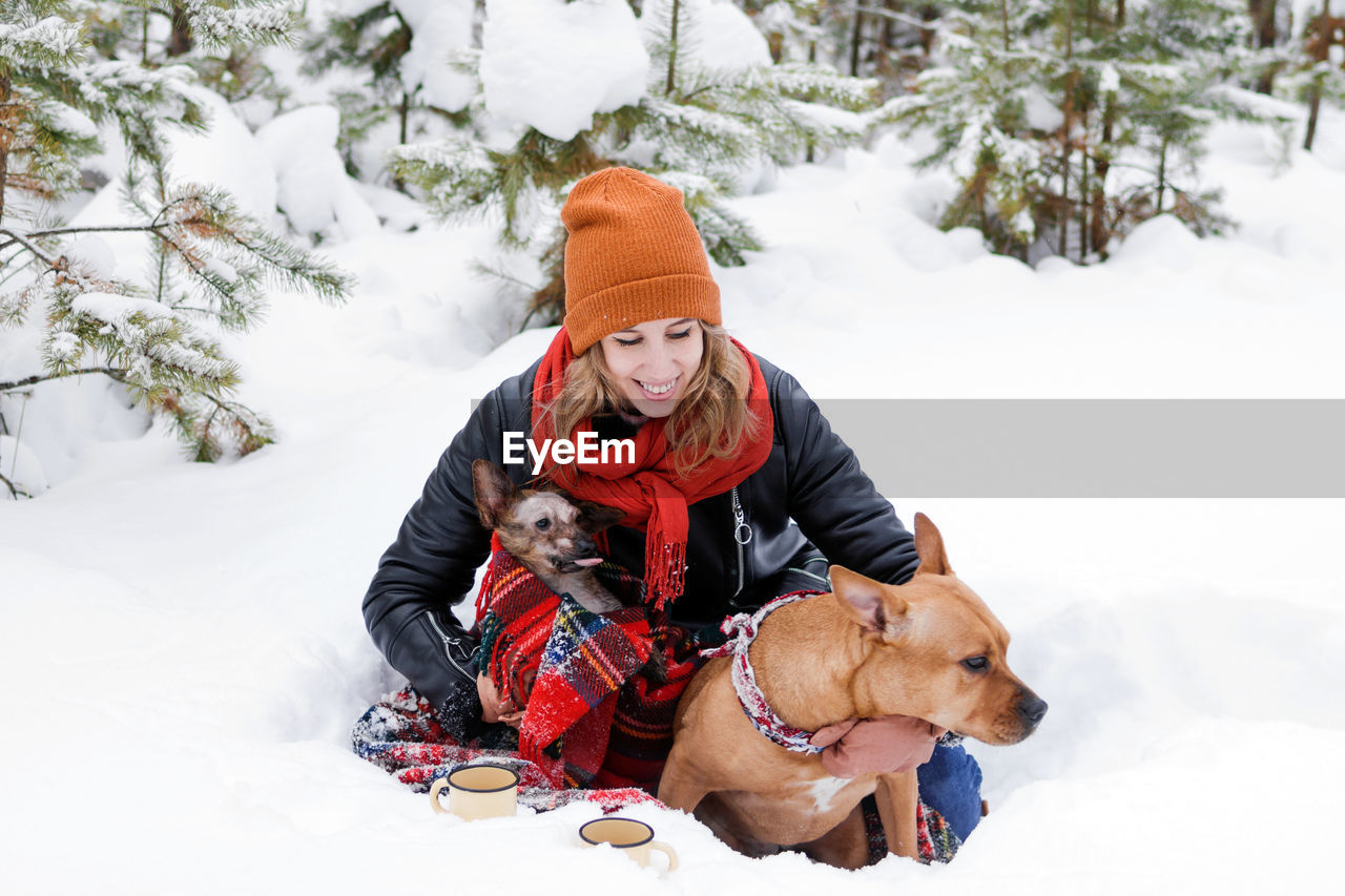Portrait of woman with her two dogs wrapped in red checkered plaid on a snow in forest.
