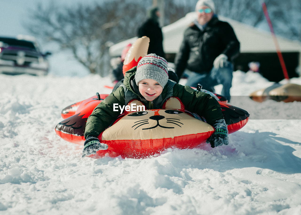 Cute boy on inflatable raft playing in snow with father in background