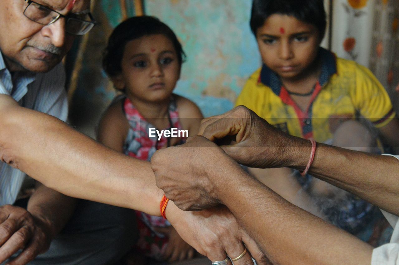 Cropped image of person tying thread to man hand while children looking at home