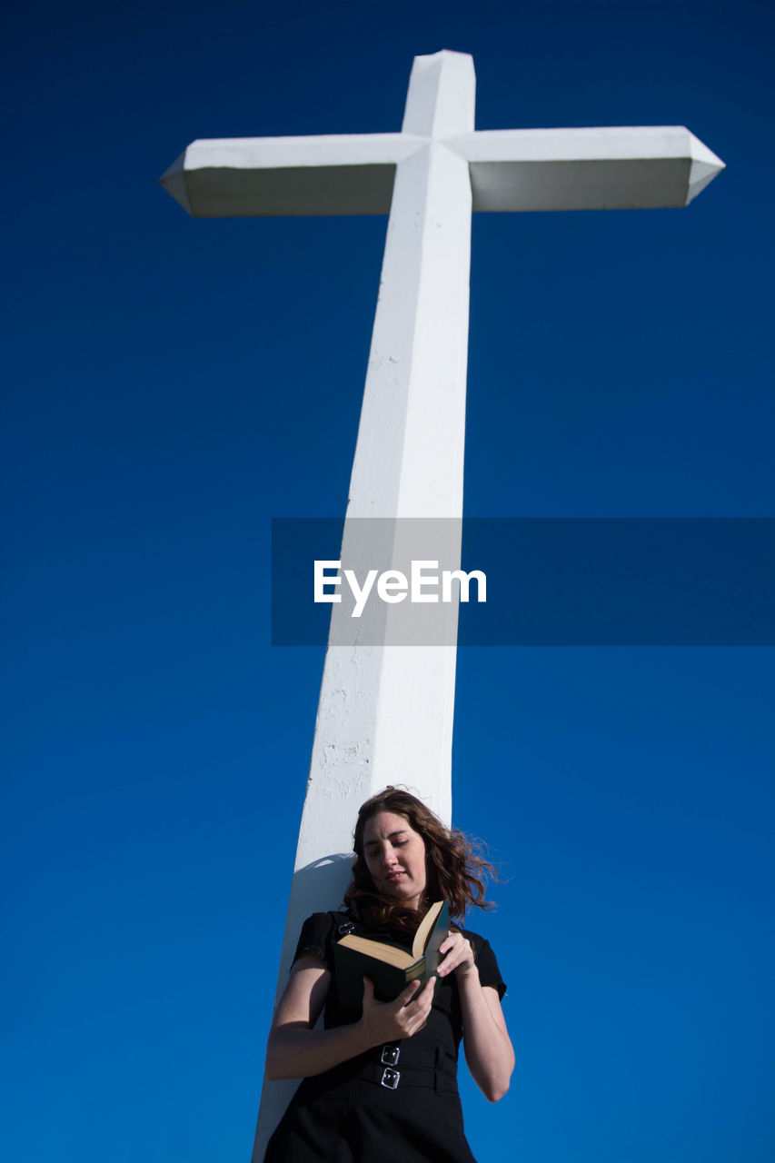 Low angle view of woman reading book while standing by religious cross against blue sky