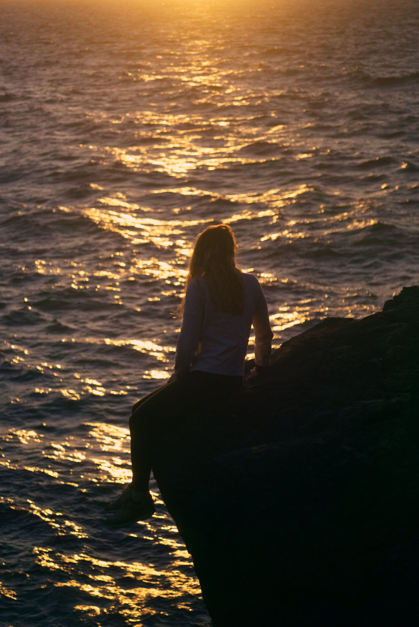Rear view of woman sitting by sea on rock during sunset