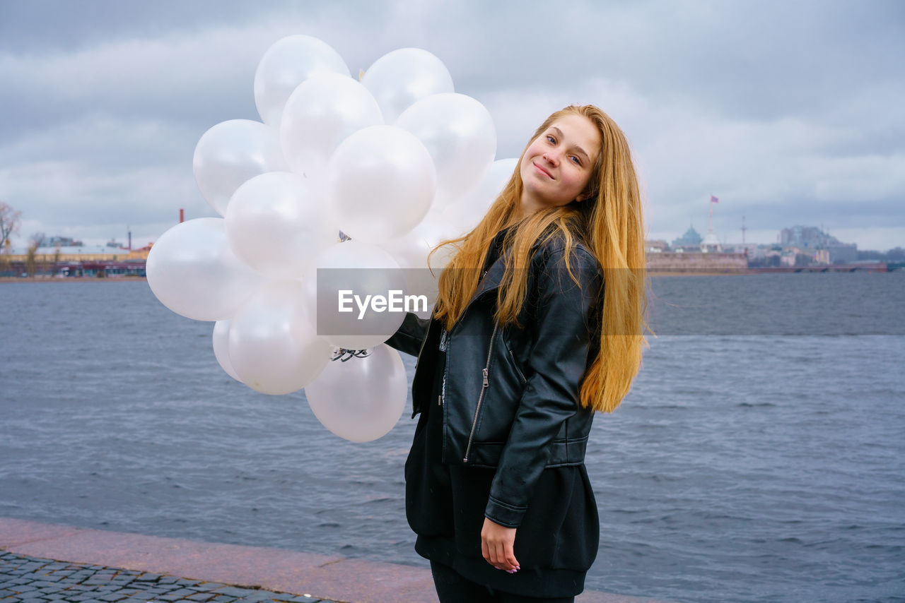 one person, long hair, young adult, water, women, sky, adult, hairstyle, cloud, nature, blond hair, emotion, smiling, happiness, standing, balloon, city, blue, clothing, three quarter length, portrait, sea, architecture, positive emotion, dress, day, outdoors, casual clothing, lifestyles, photo shoot, carefree, front view, fun, leisure activity, fashion, spring, cheerful, enjoyment, looking at camera, wind, beauty in nature, looking, smile, teeth, holiday, jacket, waist up, city life, person