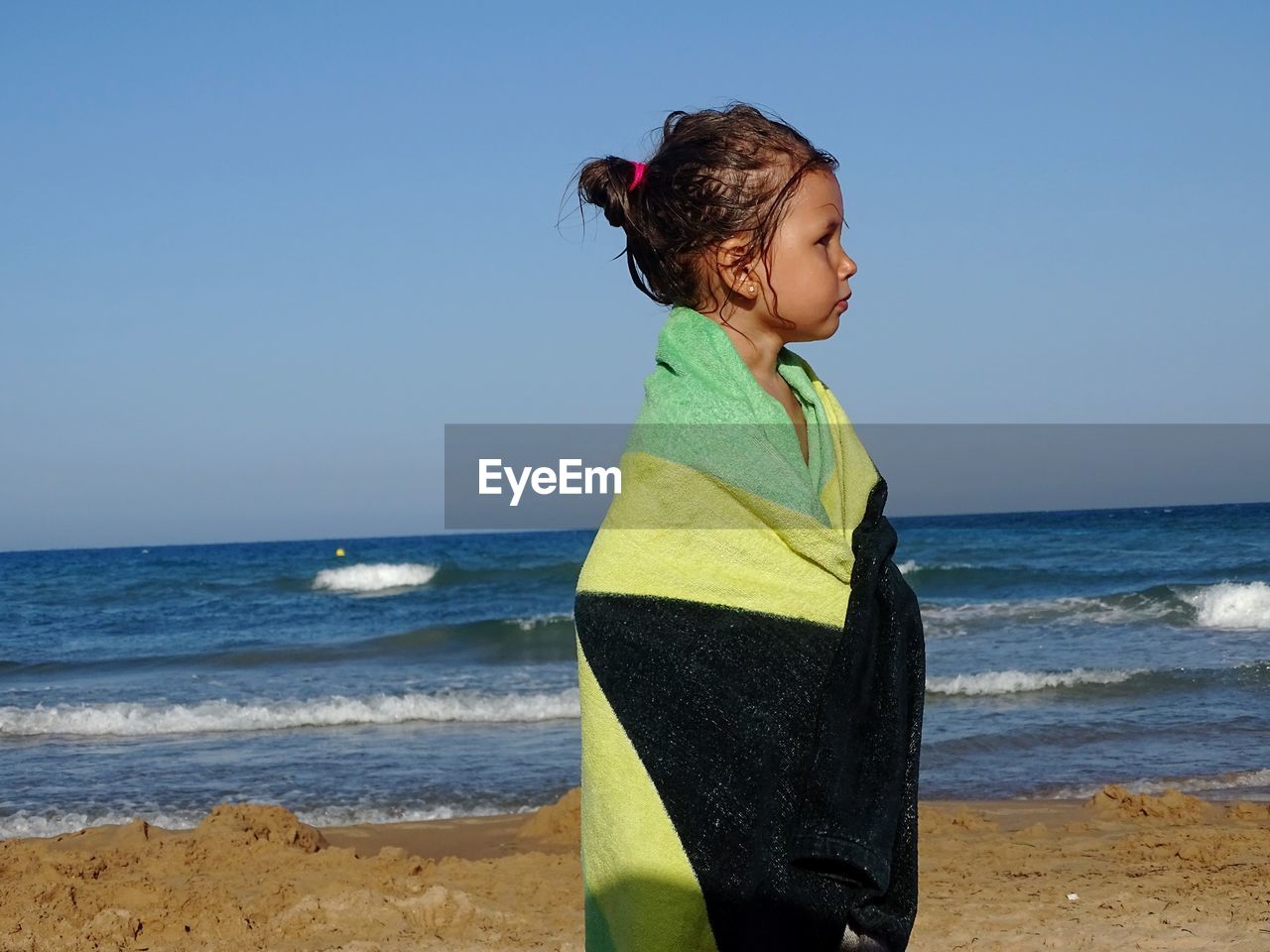 Cute girl wrapped in towel looking away while standing at beach against clear sky